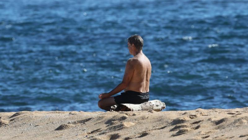 Pierce Brosnan, complet ud, în poziția lotus, meditând în fața oceanului