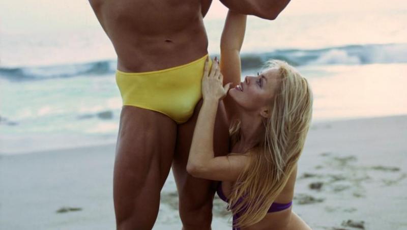 Arnold Schwarzenegger, în timpul unei ședințe foto pe malul mării, îmbrăcat într-un slip galben, având alături un model în costum de baie