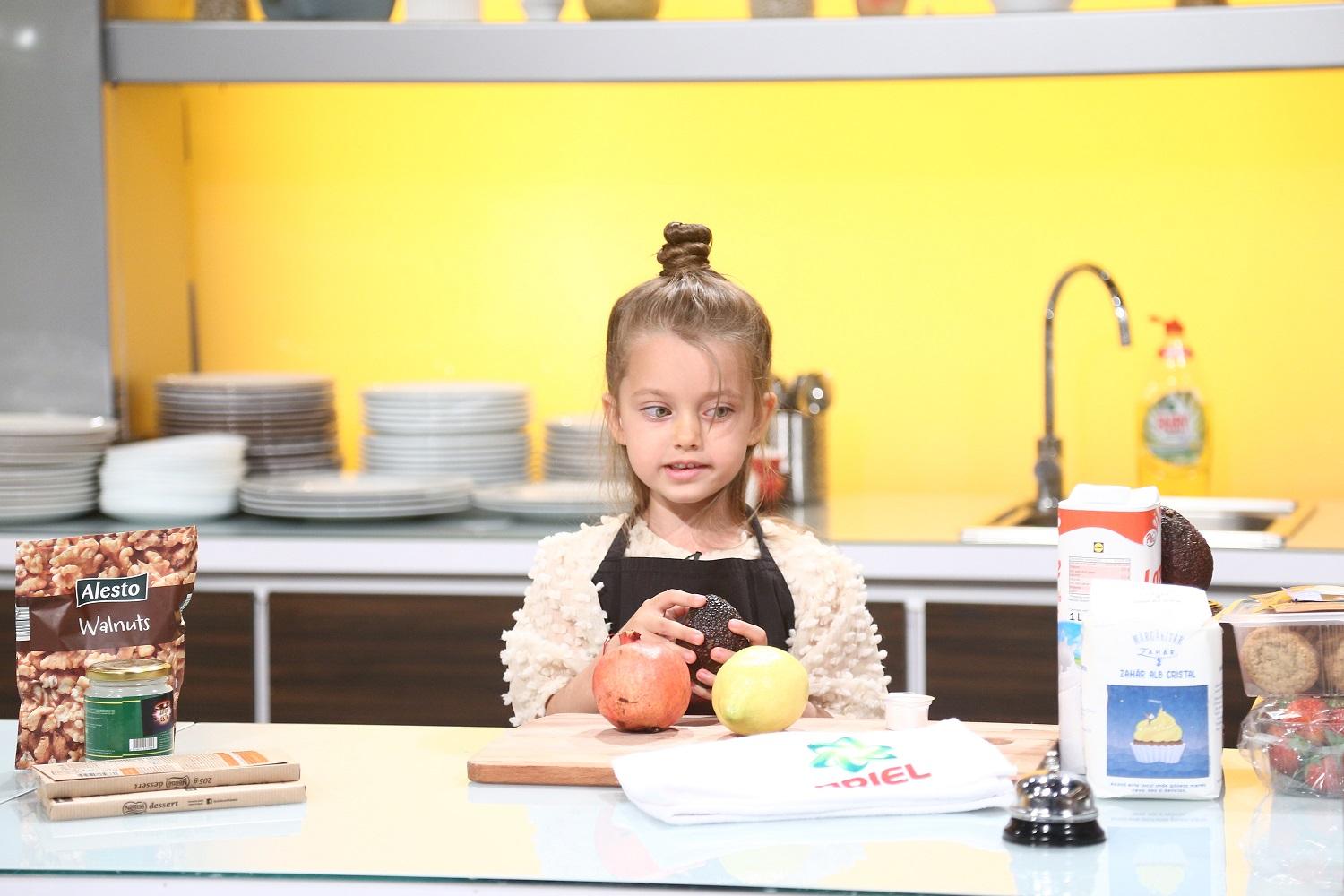 Anastasia în bucătarie, imbrăcată cu o bluză bej și un șorț negru, se pregătește să gătească, având pe masă fructe, făină și lapte.