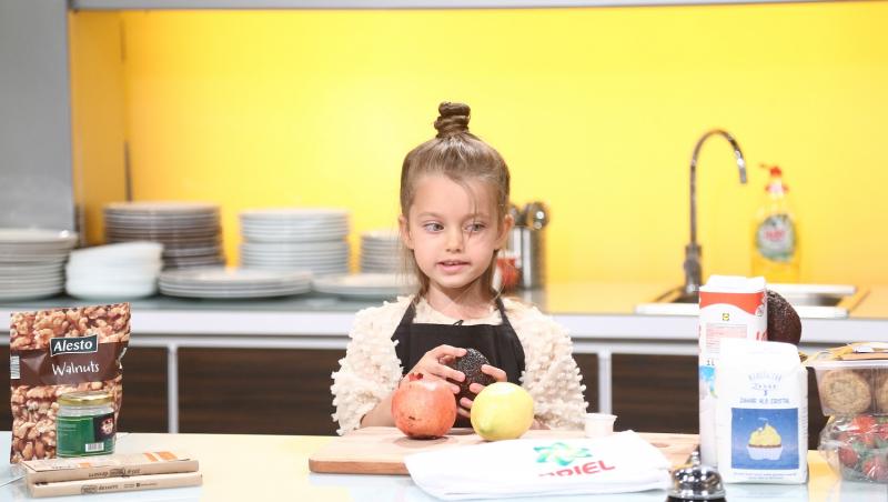 Anastasia în bucătarie, imbrăcată cu o bluză bej și un șorț negru, se pregătește să gătească, având pe masă fructe, făină și lapte.