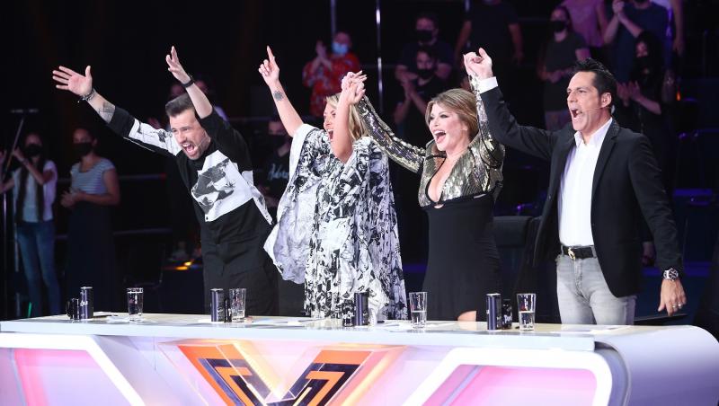 Concurenții X Factori se ridică în picioare cu mâinile sus, toti sunt imbracati elegant si par fericiti.