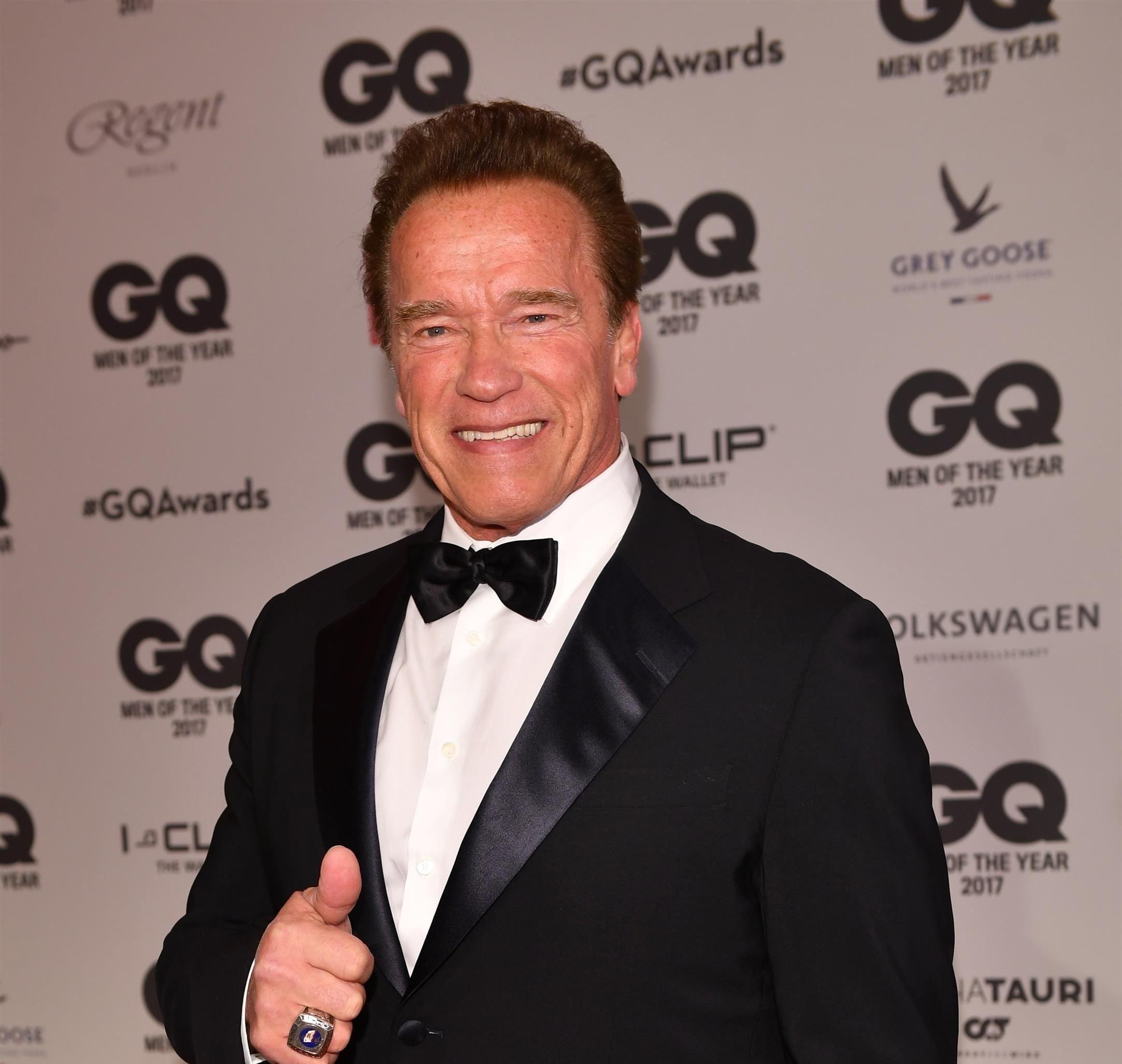 Arnold Schwarzenegger imbracat intr-un costum negru, cu camasa alba si papion negru. Zambeste, se uita la camera si isi arata degetul mare.