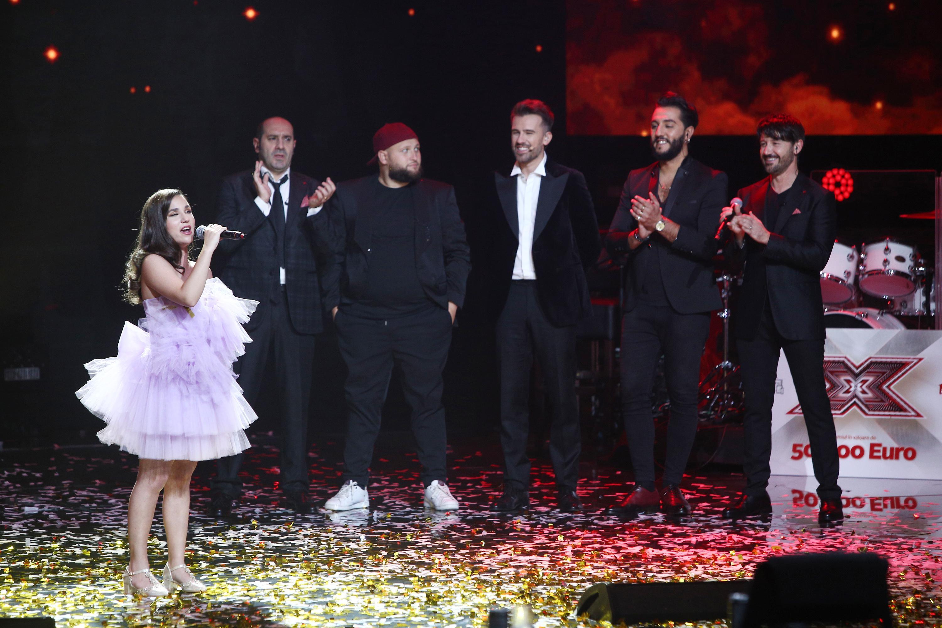 Andrada Precup, într-o rochie lila cu multe volane, cântă pe scena X Factor, în timp ce italienii de la Super 4 o privesc cu admirație