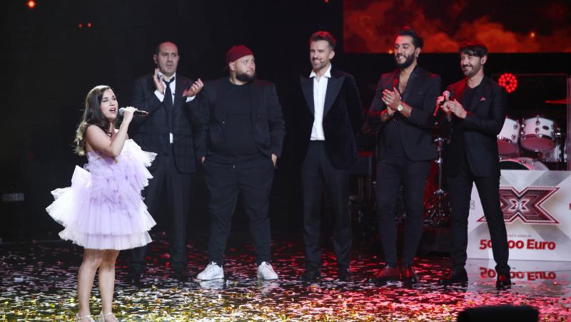 Andrada Precup, într-o rochie lila cu multe volane, cântă pe scena X Factor, în timp ce italienii de la Super 4 o privesc cu admirație