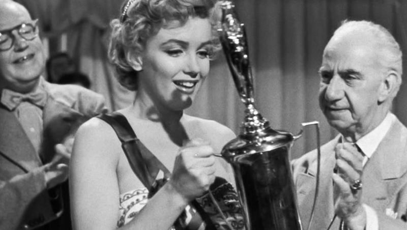 Marilyn Monroe înconjurată de barbati, tinand un premiu in maini