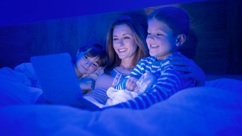 mama cu doi copii in pat uitandu-se la laptop pe intuneric
