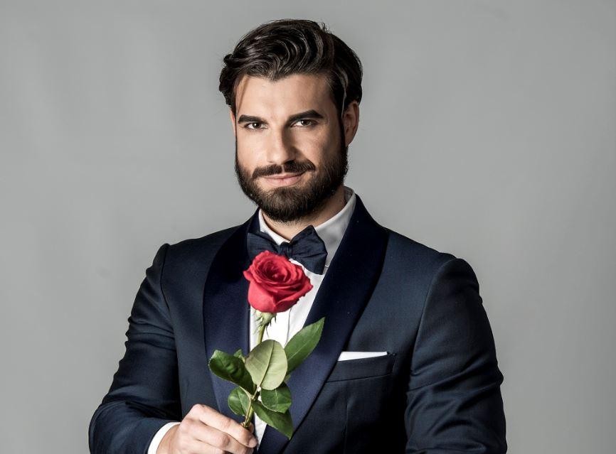 Andi Constantin, burlacul sezonului 6 al show-ului, îmbrăcat la costum, cu papion, ține în mână un trandafir roșu și zâmbește mistrios