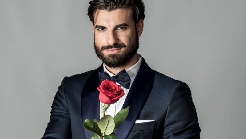 Andi Constantin, burlacul sezonului 6 al show-ului, îmbrăcat la costum, cu papion, ține în mână un trandafir roșu și zâmbește mistrios