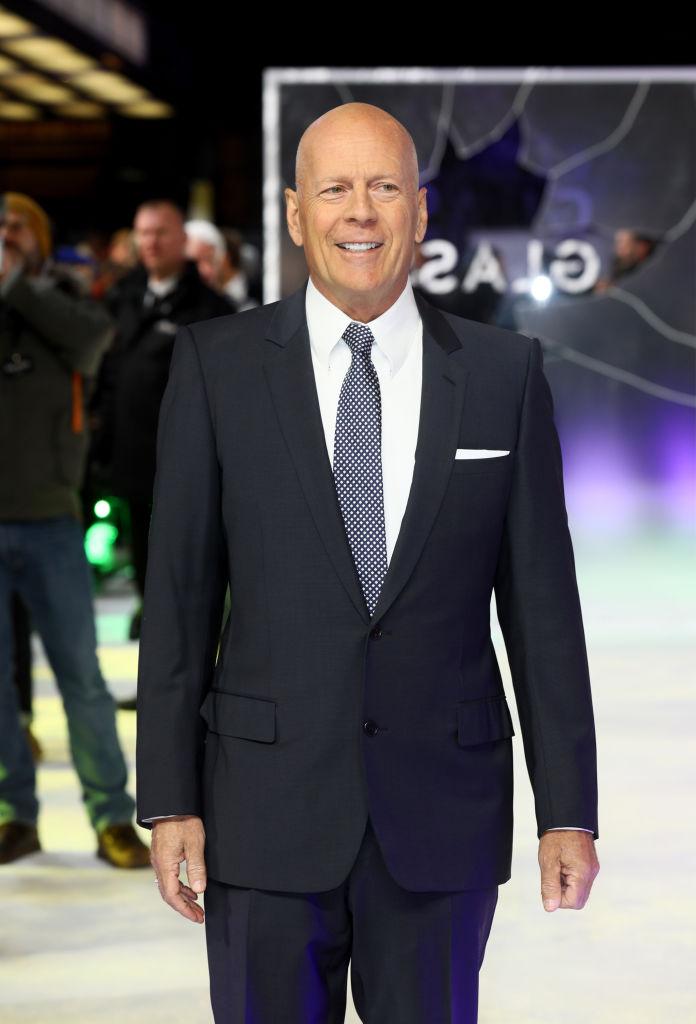 Bruce Willis pe covorul rosu intr-un costum negru si camasa alba