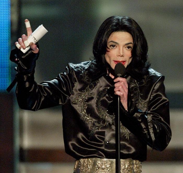 Michael Jackson intr-un costum negru, vorbeste la microfon si tine un deget sus