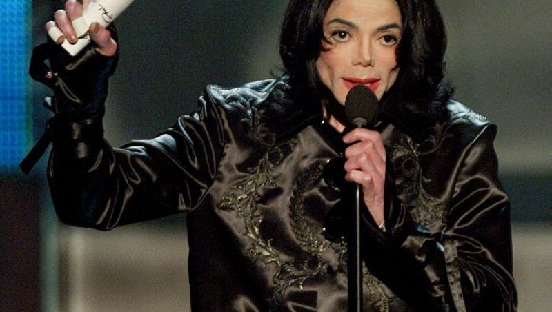Michael Jackson intr-un costum negru, vorbeste la microfon si tine un deget sus
