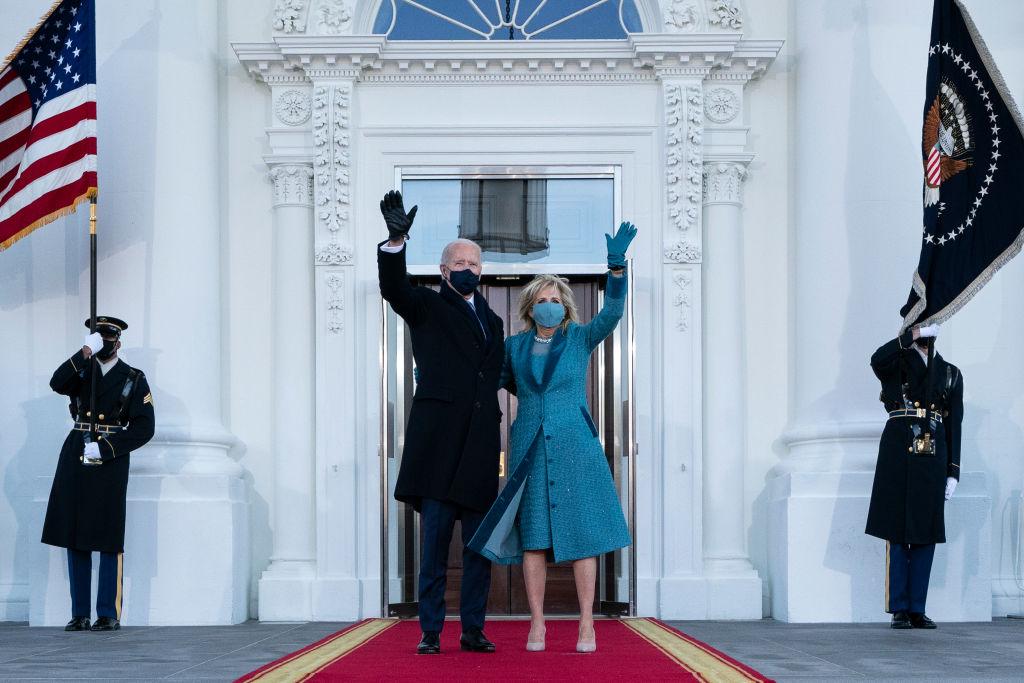 Joe Biden, îmbrăcat la costum și Jill Biden, cu un palton albastru, în fața unor uși închise, la Casa Albă, făcând cu mâna populației