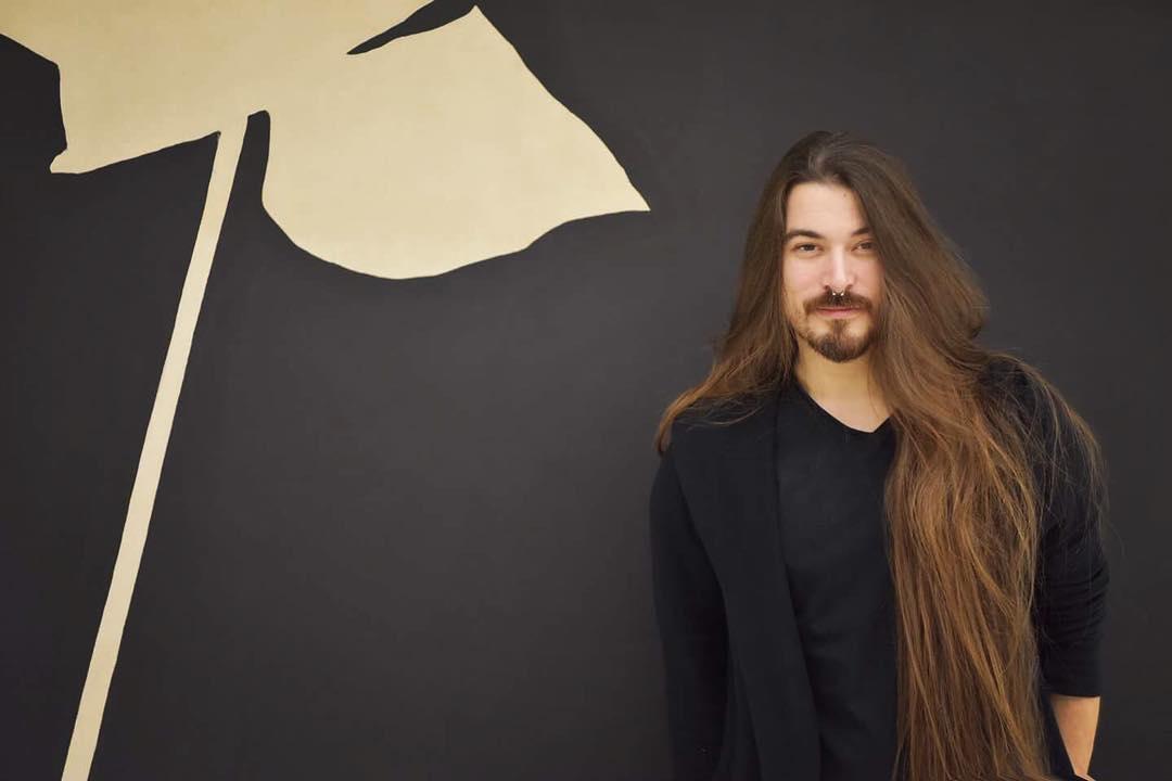 Massimo Volpe, cu părul despletit, făcând o poză cu spatele la un perete cu element decorativ