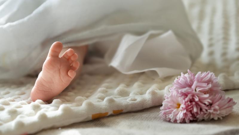 picior de bebelus pe o paturica alba, infasat in cearceaf alb si o floare mov langa