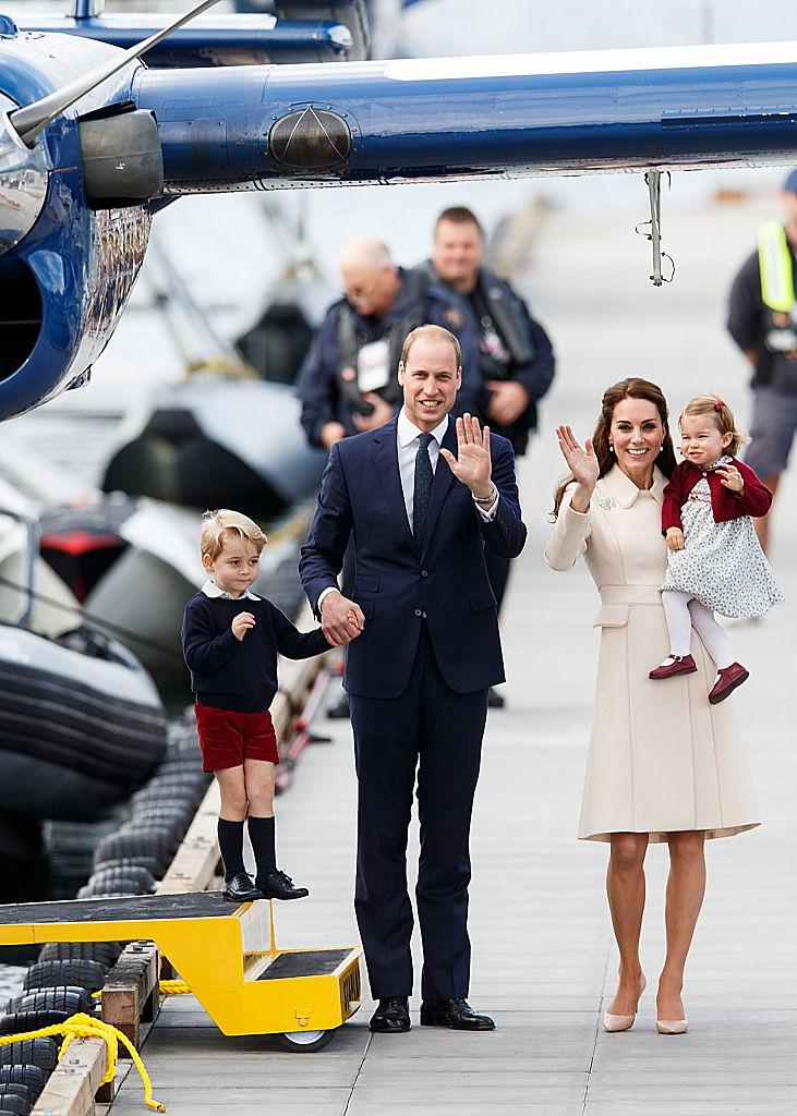 Printul William imbracat intr-un costum negru paseste alaturi de Kate Middleton imbracata cu un trench alb paseste alturi de cei doi copii in aeroport si fac cu mana la camera