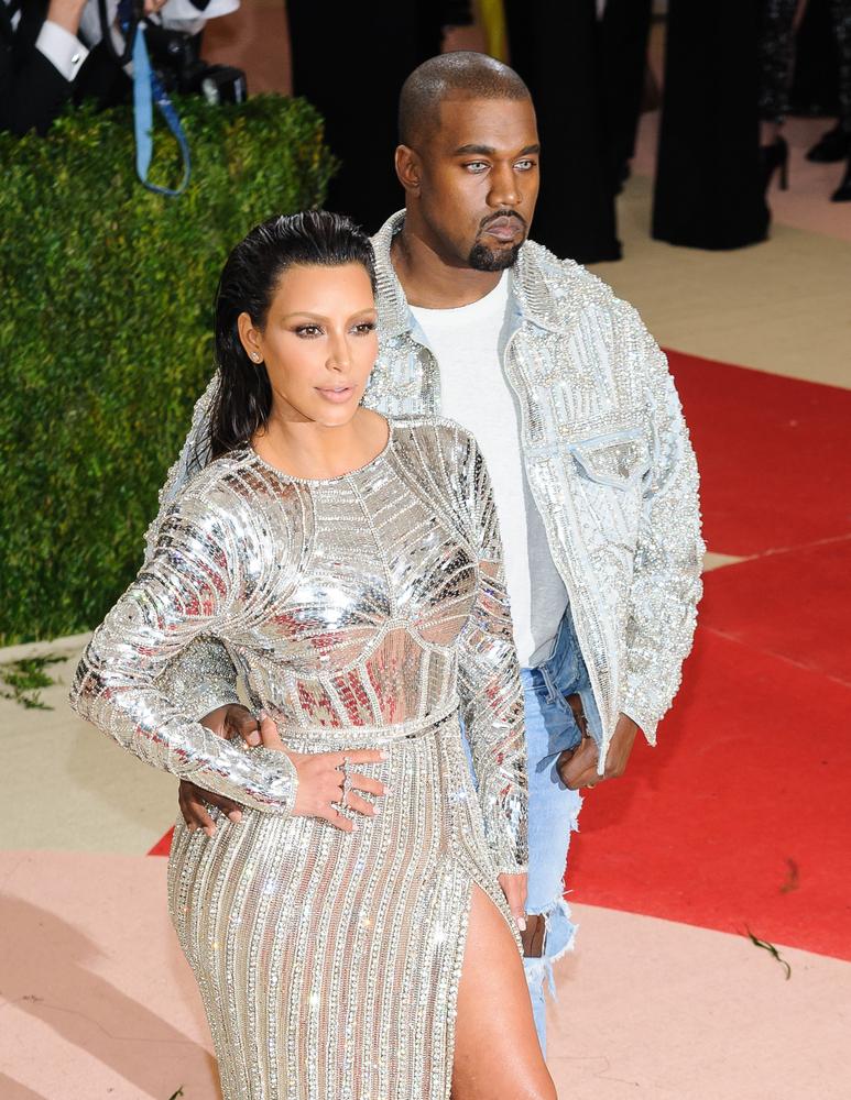 Kanye West imbracat cu un sacou gri si o bluza alba alaturi de Kim Kardashian care poarta o rochie eleganta argintie