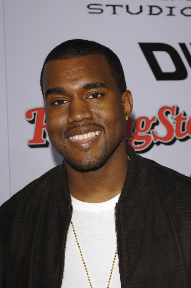 Kanye West imbracat intr-un sacou negru si un tricou alb se uita spre camera de fotografiat si zambeste