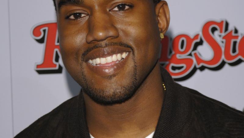 Kanye West imbracat intr-un sacou negru si un tricou alb se uita spre camera de fotografiat si zambeste