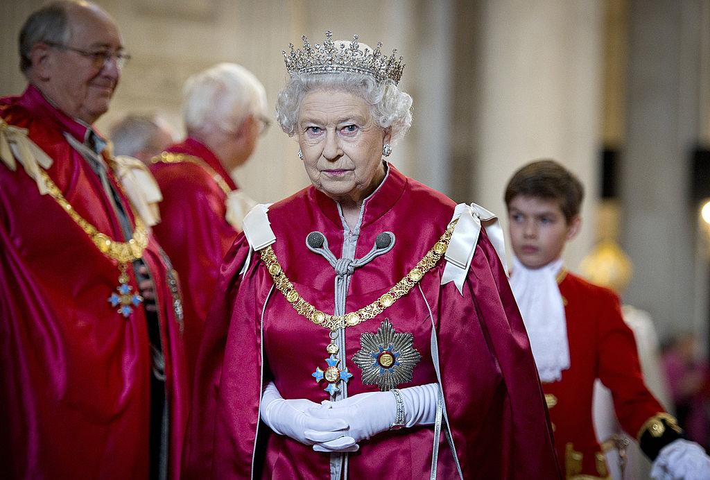 Regina Elisabeta a Marii Britanii imbracată într-o capă roșie și mănuși albe si purtand pe cap o coroana cu pietre pretioase