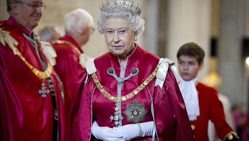 Regina Elisabeta a Marii Britanii imbracată într-o capă roșie și mănuși albe si purtand pe cap o coroana cu pietre pretioase
