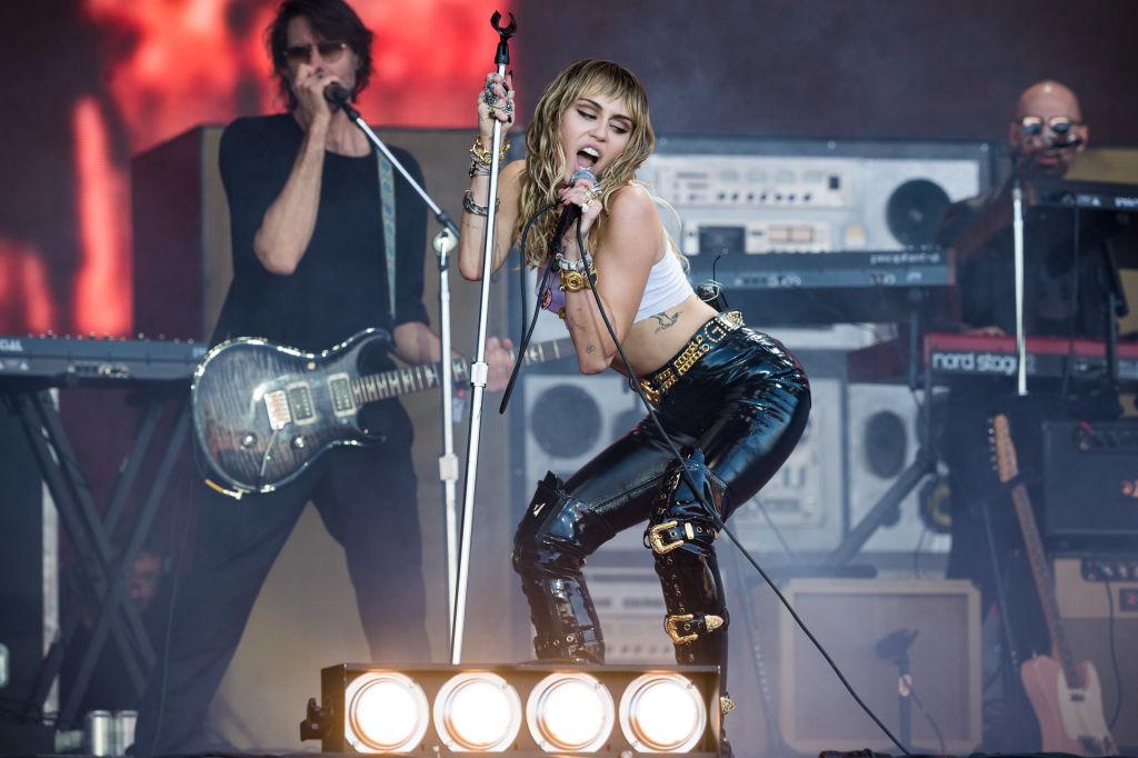 Miley Cyrus, cu părul lung, purtând o pereche de pantaloni de piele negri, pe scenă