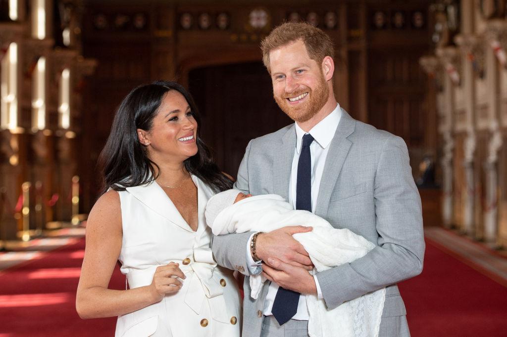 Meghan Markle într-o rochie albă și Prinţul Harry într-un costum gri, ținându-l în brațe pe fiul lor Archie