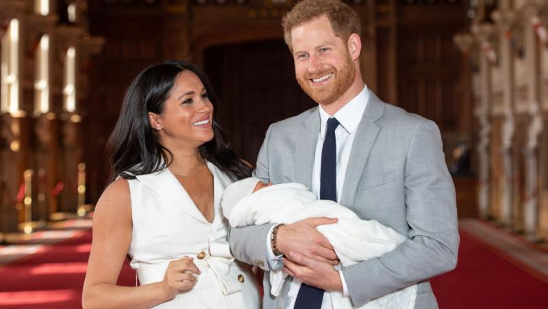Meghan Markle într-o rochie albă și Prinţul Harry într-un costum gri, ținându-l în brațe pe fiul lor Archie