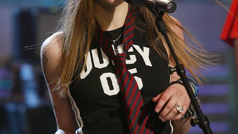 Avril Lavigne intr-un tricou negru, pantaloni gri, are o cravată vișinie și cântă la microfon