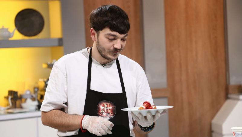 Theo Costache, cu șorțul Chefi la cuțite, este la masa de gătit și ține o farfurie într-o mână