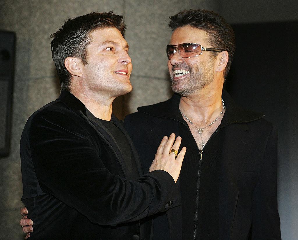 George Michael și fostul lui iubit, Kenny Goss, îmbrăcați amândoi cu sacouri negre. George Michael zâmbește către Goss