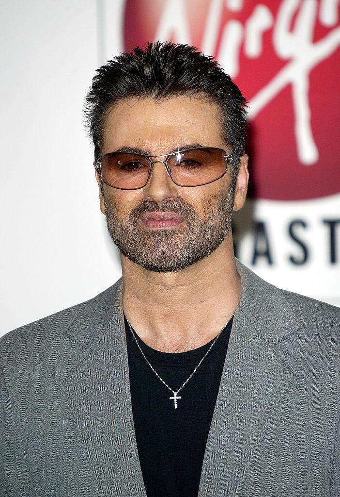 George Michael, îmbrăcat în sacou gri cu o bluză neagră pe dedesubt, și ochelari