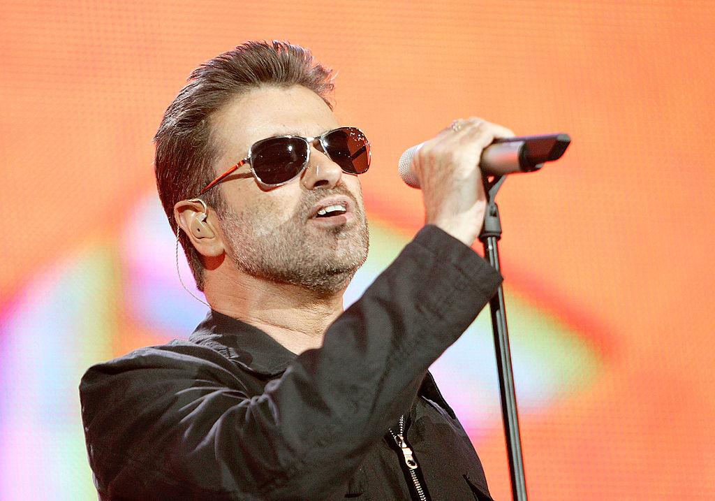 George Michael, îmbrăcat în negru, cu ochelari la ochi, cântă la microfon