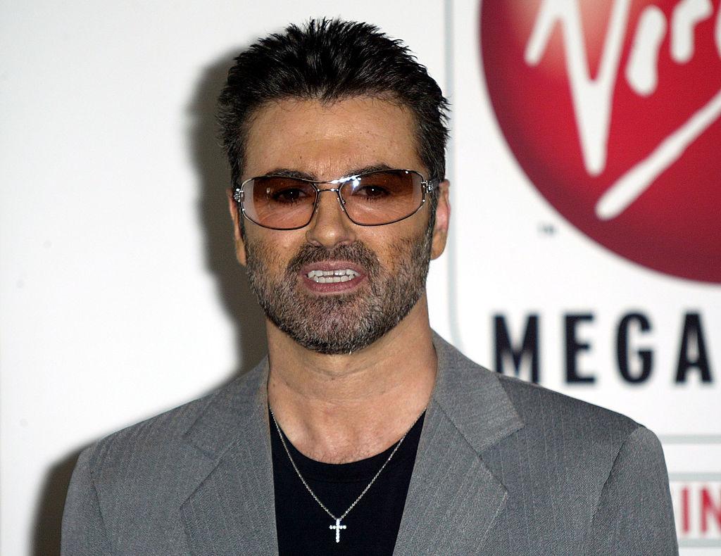 George Michael, îmbrăcat în sacou gri cu o bluză neagră pe dedesubt, și ochelari