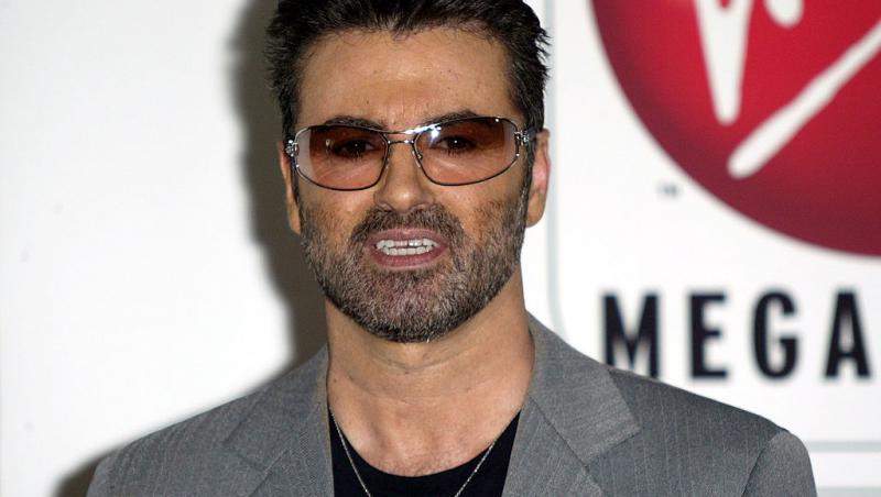 George Michael, îmbrăcat în sacou gri cu o bluză neagră pe dedesubt, și ochelari