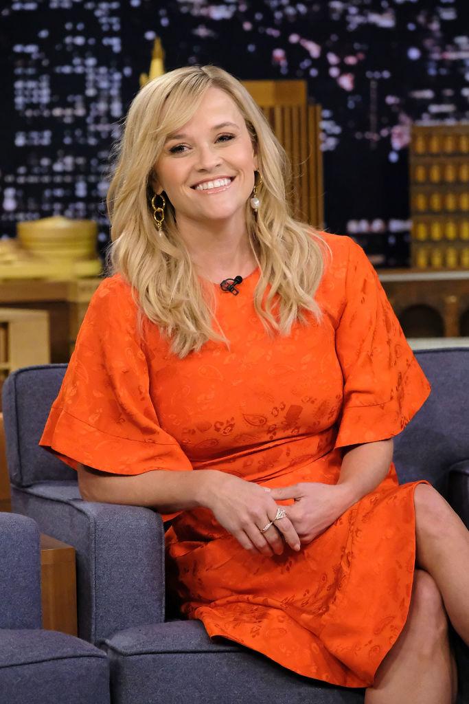 Reese Witherspoon îmbrăcată într-o rochie portocalie, cu părul desfăcut, râde