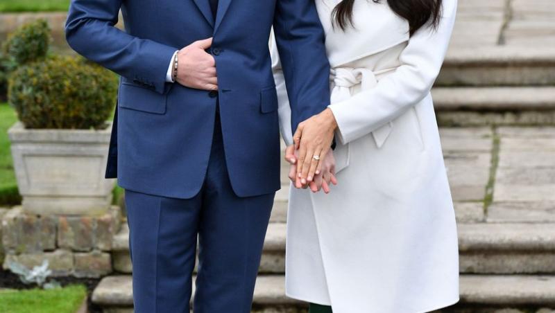 Prințul Harry și Meghan Markle, îmbrăcați elegant. El este într-un costum albastru, ea în rochie și palton deschis la culoare
