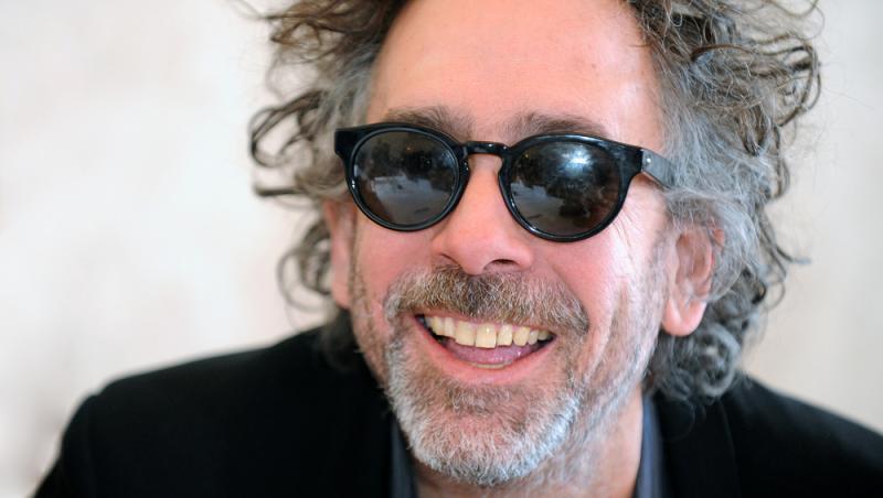 Tim Burton zâmbește, are ochelari de soare și este îmbrăcat în negru