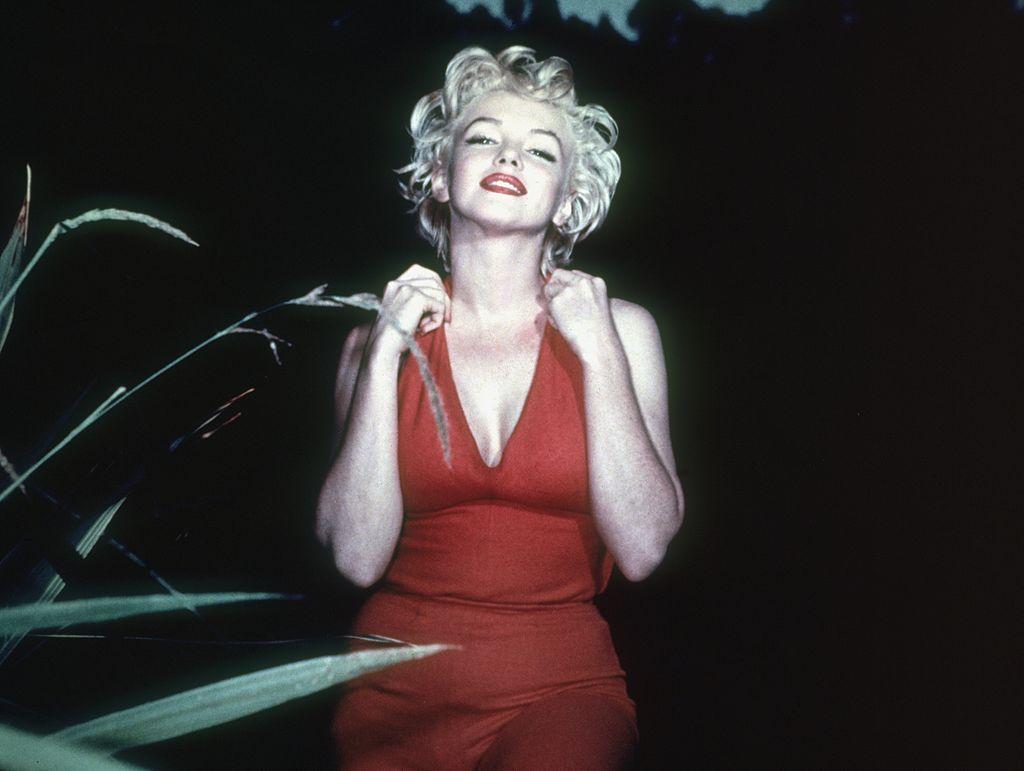 Marilyn Monroe, zâmbitoare, îmbrăcată într-o rochie roșie, își ine mâinile în preajma gâtului