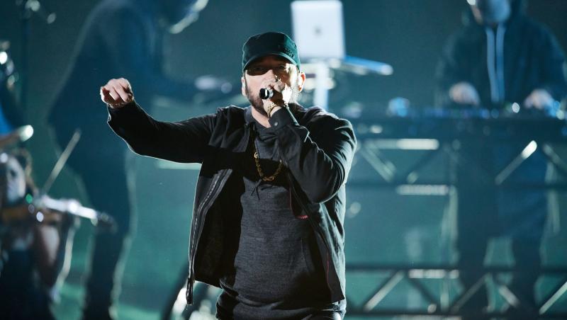 Eminem, îmbrăcat în haine închise la culoare, pe scenă, cântând