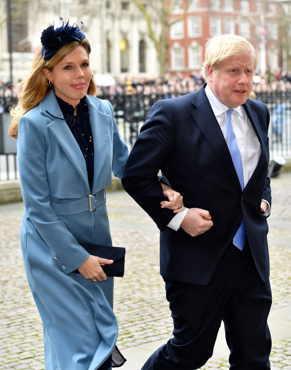 Carrie Symonds, soția lui Boris Johnson, purtând un costum albastru, ținându-l d emână pe premierul britanic, îmbrăcat în sacou și pantaloni de stofă