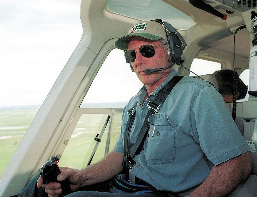 Harrison Ford, la bordul unei aeronave, cu ochelari la ochi și șapcă, îmbrăcat într-o bluză albastră
