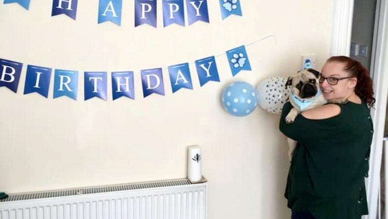janice kerwick cu o bluza neagra, tinand in brate cainele ei si pe perete scrie happy birthday