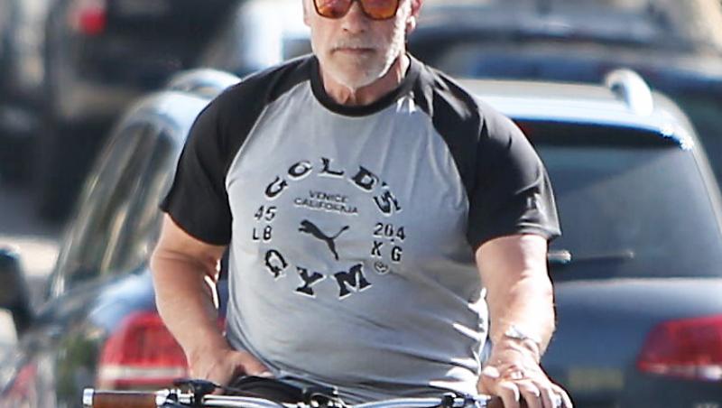 Schwarzenegger purta o șapcă și ochelari de soare, pantaloni scurți și un tricou cu inscripția numelui unei săli de forță