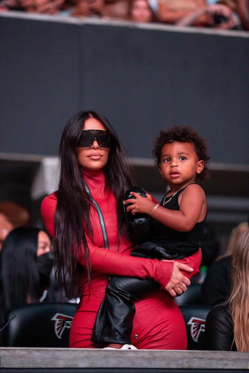kim kardashian imbracata in rosu, cu ochelarii de soare negri la ochi, tinandul in brate pe fiul ei, la lansarea lui kanye west