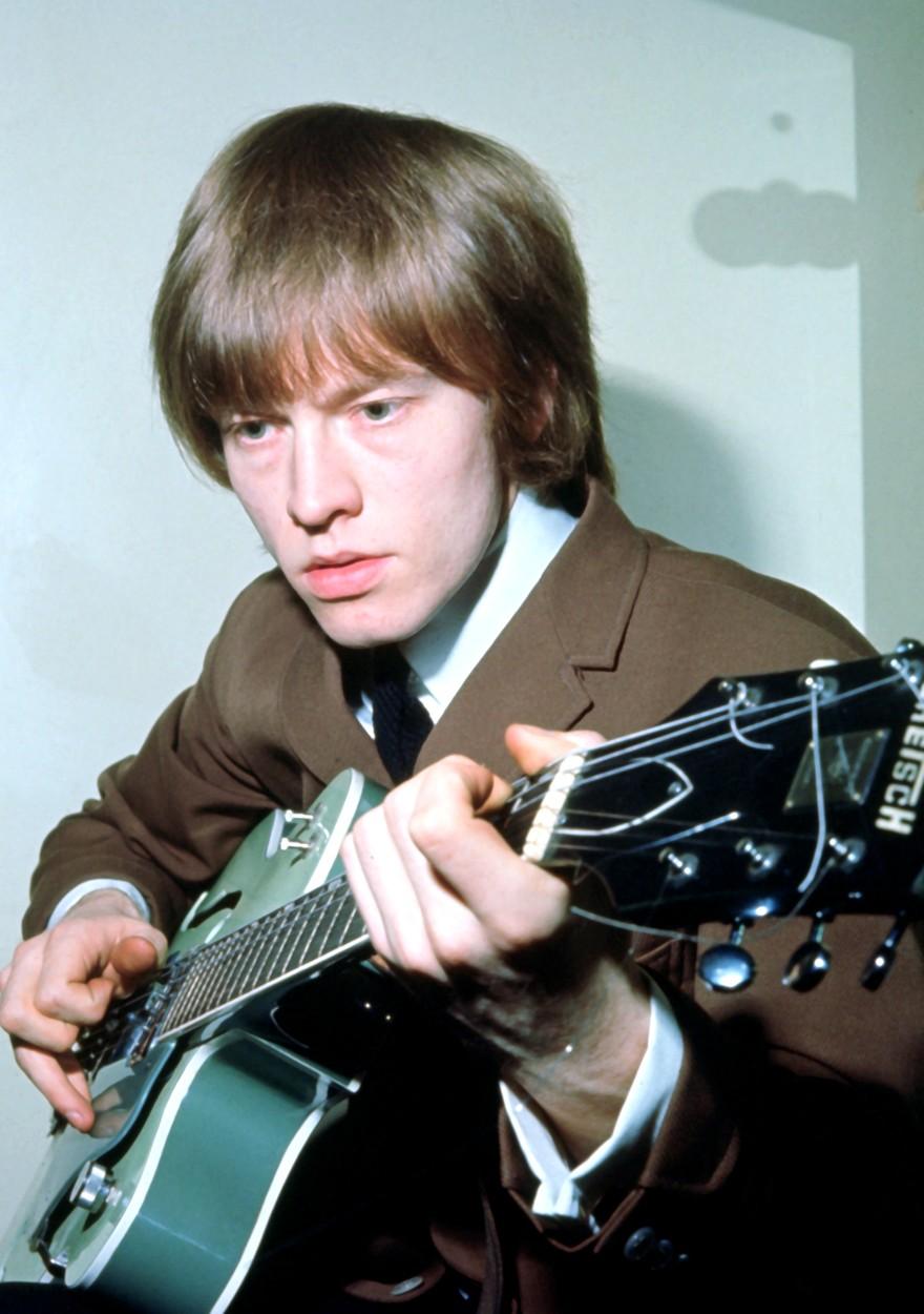 brian jones intr-o camasa alba cu sacou kaki, cu chitara in mana