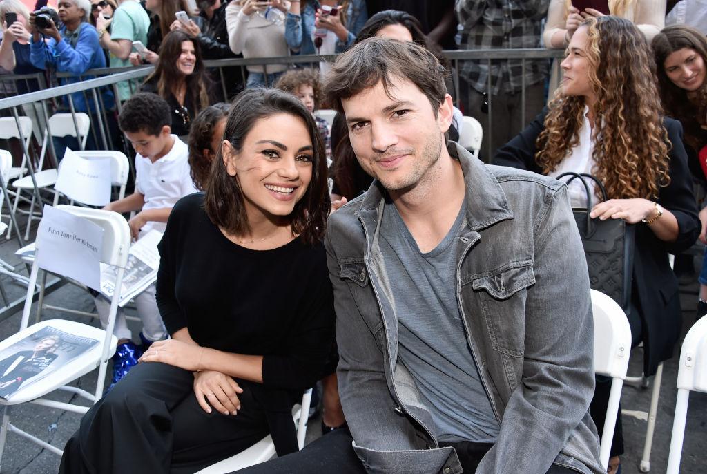 Mila Kunis și Ashton Kutcher așezați pe niște scaune albe, îmbrăcați casual