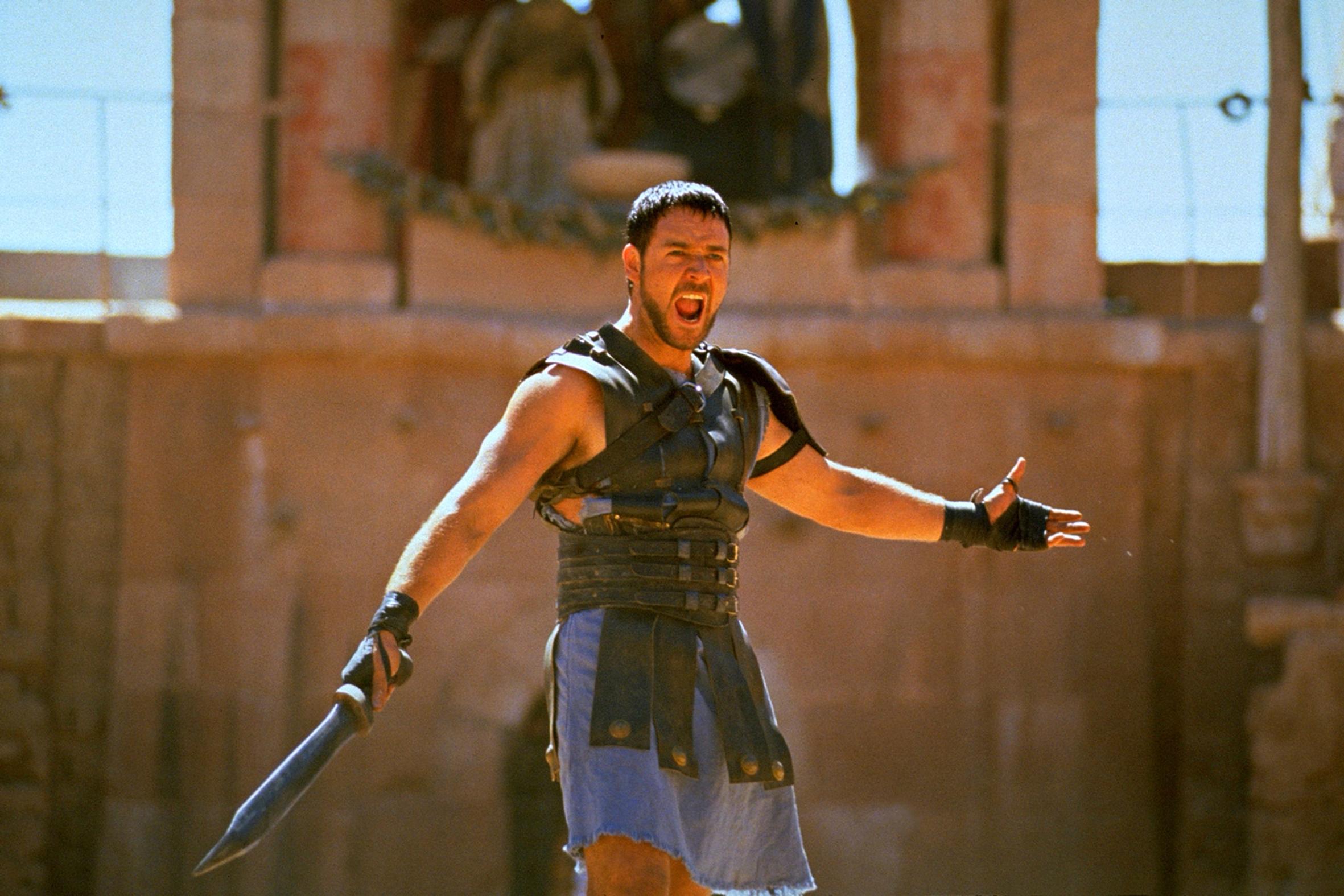 russell crowe în filmul gladiatorul, țipând la public, în arena gladiatorilor. are sabia în mână și e îmbrăcat în armură, cu fundal bej în spate