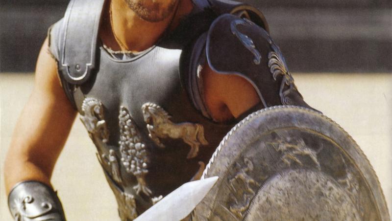 russell crowe în filmul gladiatorul, în arena gladiatorilor. are sabia în mână și e îmbrăcat în armură, se apără cu scutul, cu fundal bej în spate