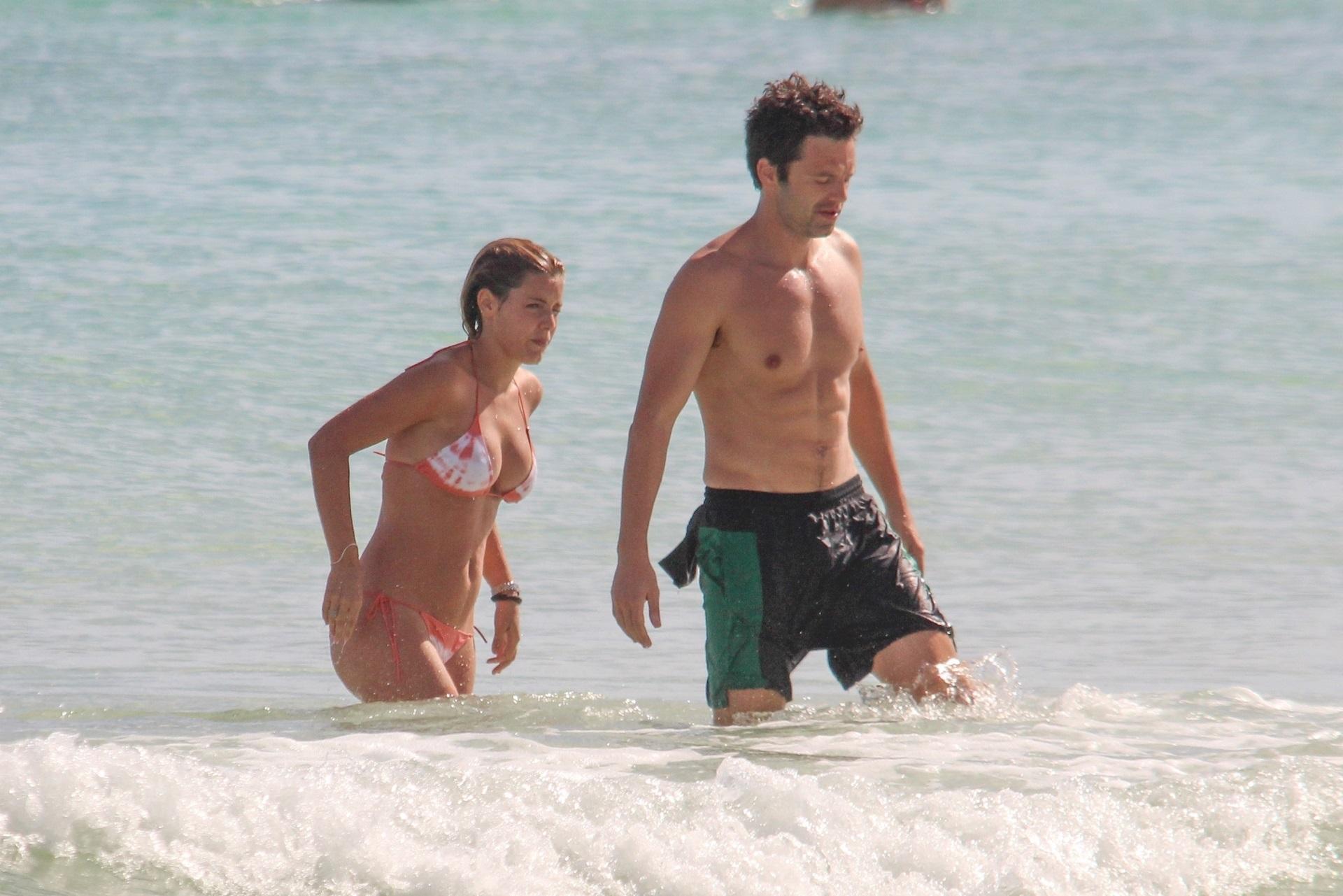 sebastian stan și alejandra onieva la plajă. fundal cu mare, ei ies din valuri. ea poartă un bikini cu alb și portocaliu, el poartă un costum de baie cu negru și verde