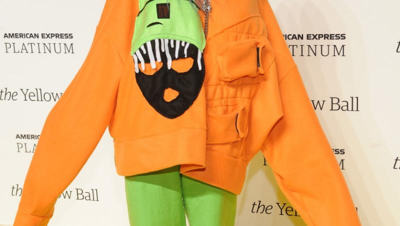 billie eilish intr-o camasa portocalie cu maneca lunga, pantaloni verzi si tenesi verzi